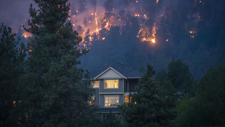A two-story home with lights on at dusk in the foreground, with active flames burning across the forested hillside directly above — a wildfire approaching a residential property in western Canada.