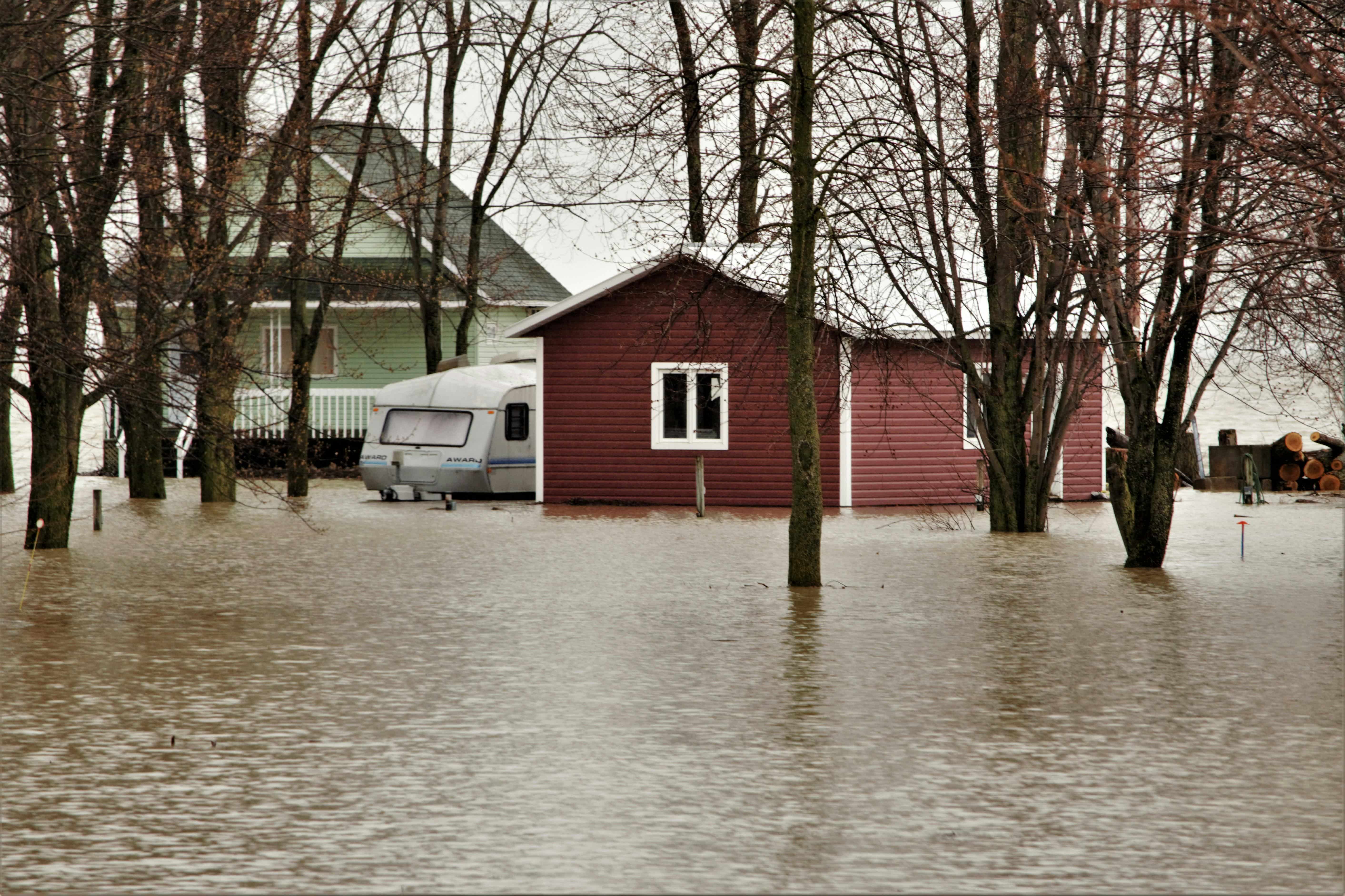 A red shed and white trailer surrounded by muddy brown floodwater and bare trees, with a green house in the background — a residential property during spring flooding in Canada.